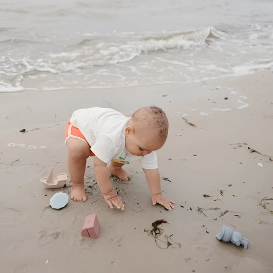 Ein baby in weißem shirt und orangen shorts krabbelt am wasserstrand neben schimmelresistentem badespielzeug piraten
