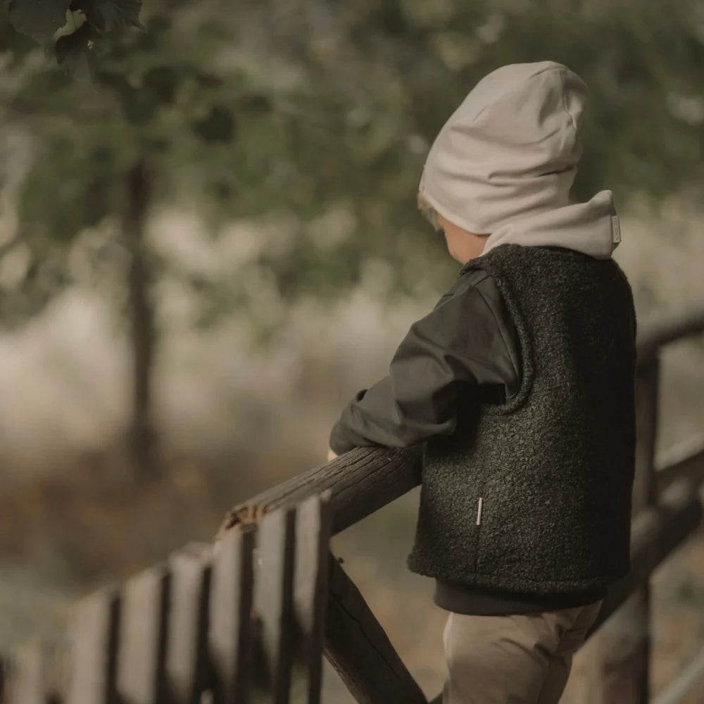 Kind eines Kindes in beigen Beanie und dunkler Weste lehnt an Holzgeländer