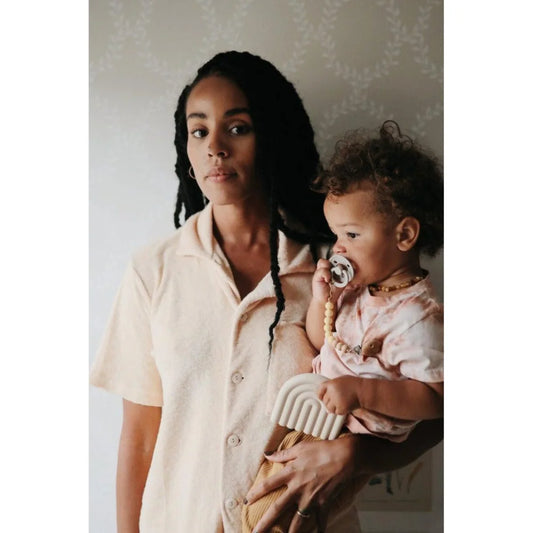 Frau hält ein kleines Kind mit Schnuller neben Beissring Regenbogen in Sandfarbe