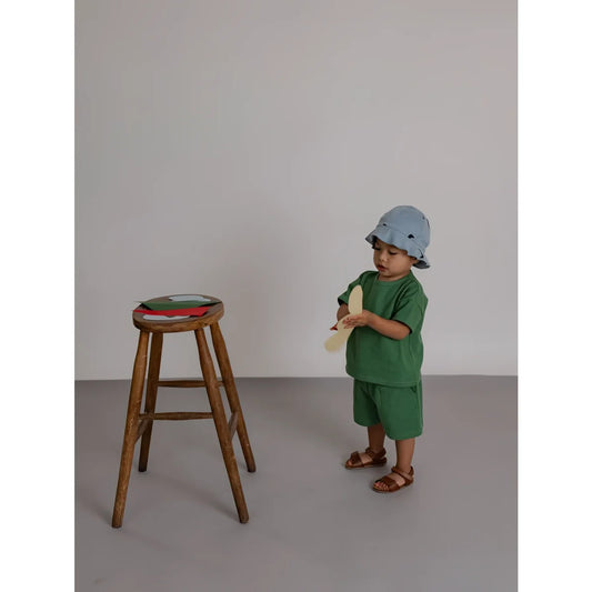 Ein junges kind in grünen bermudas aus seegras steht neben einem rustikalen hocker