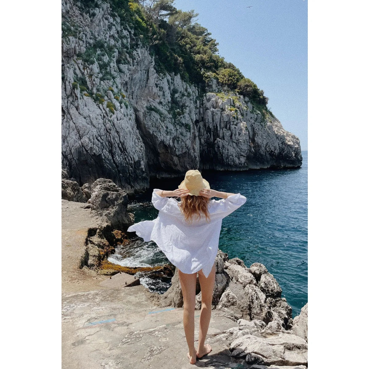 Frau in weißem Hemd und Sonnenhut steht an einem felsigen Strand. Bluse Palma | Blanco