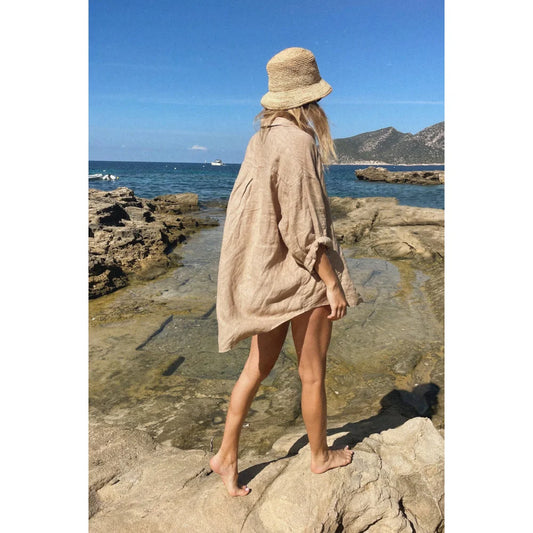 Dame mit beigem Oversized-Hemd und Strohhut auf felsigem Küstengelände