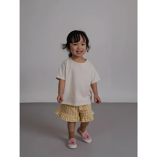 Kind mit Boxy T-Shirt Watch Me Grow, karierten Shorts und Kirschenschuhen