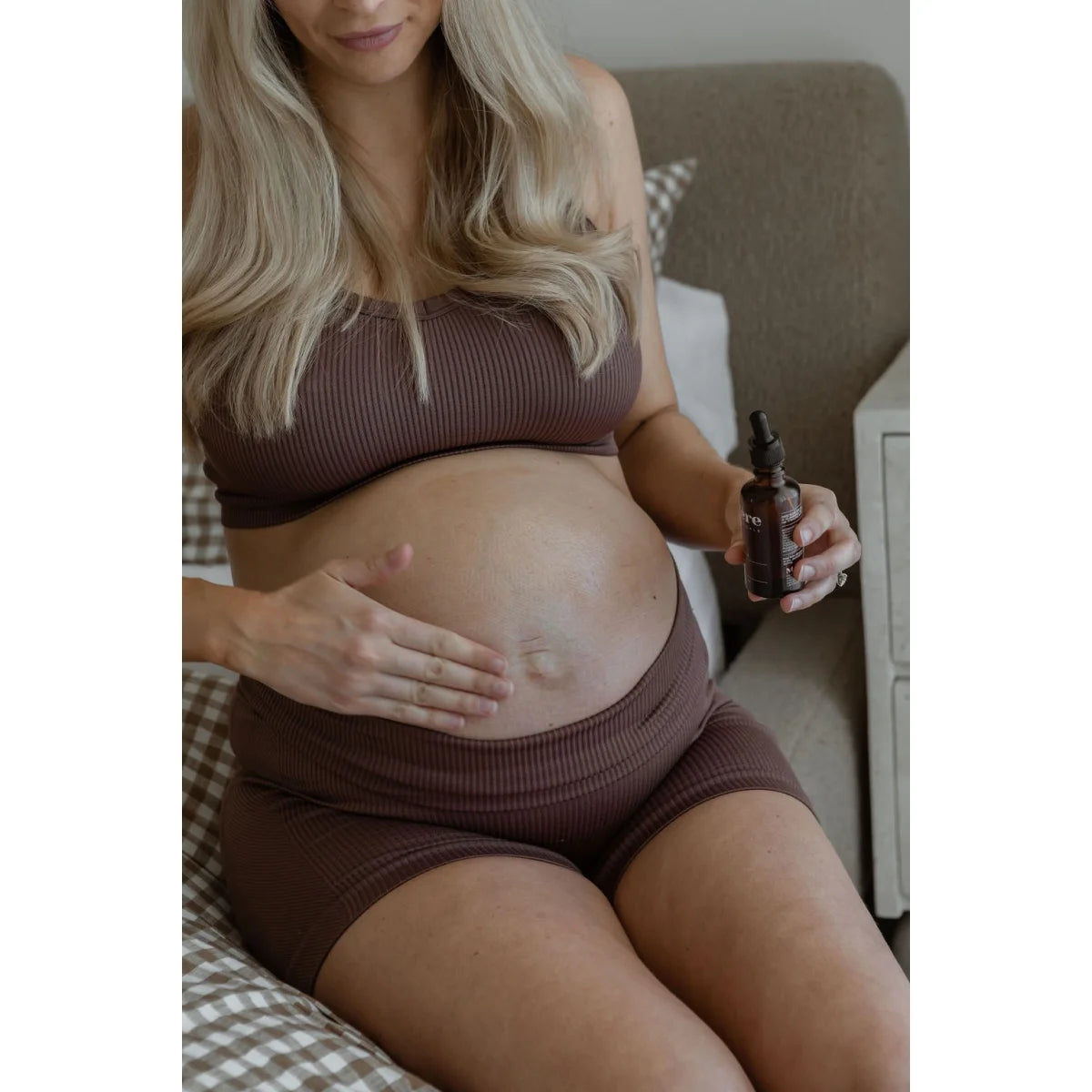 Schwangere Frau in braunem Strick-Loungewear hält ihren Bauch und eine Flasche C-Oil