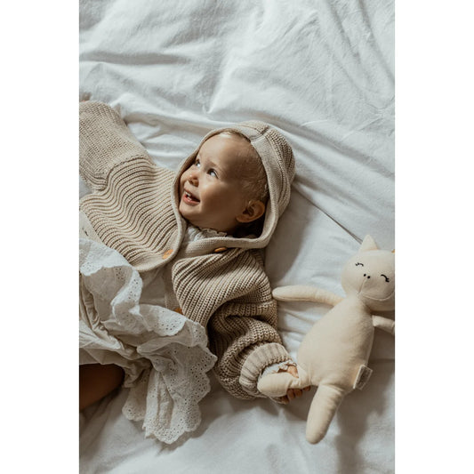 Lächelndes Baby in Strickjacke auf Bettwäsche mit Stofftier in der Nähe
