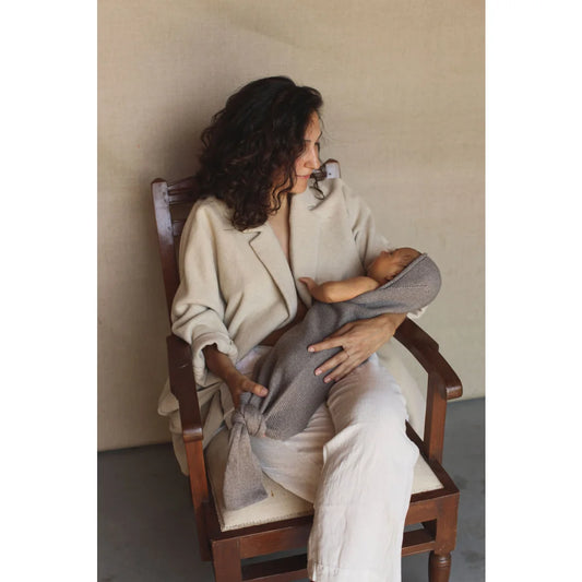 Frau mit lockigem, dunklem Haar in cremefarbenem Outfit sitzt auf Holzstuhl mit Cocoon Babydecke Otter