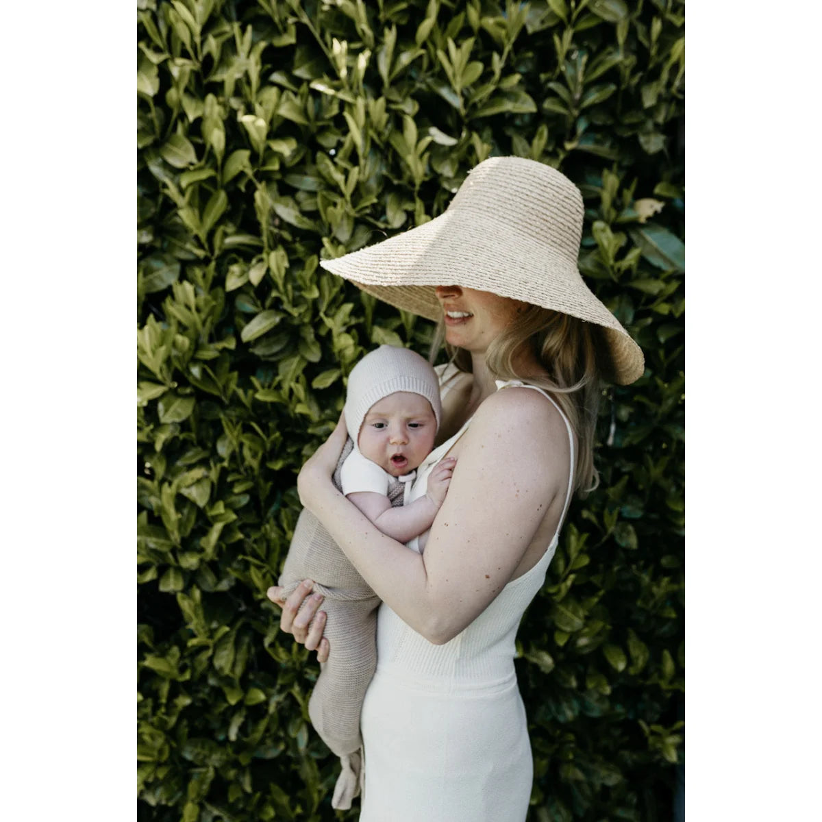 Frau mit Sonnenhut hält Baby vor blättrigem Hintergrund, präsentiert Cocoon Babydecke Sand