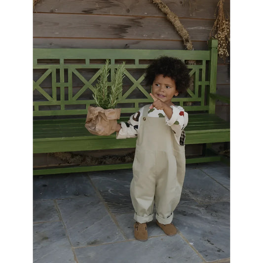 Kind im cremefarbenen Overall hält einen Blumentopf, Farmer Dungarees Latzhose von Seasalt