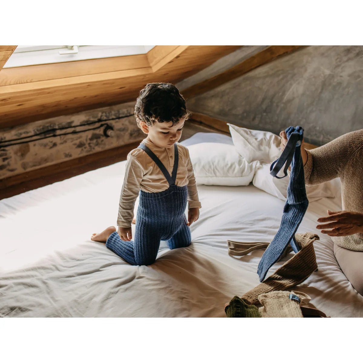 Junge im blauen gestreiften Latzhose und beigem Shirt, kniet auf einem Bett