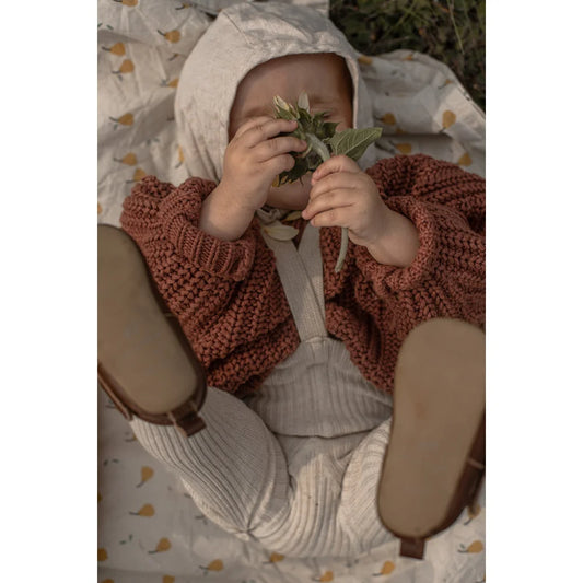Baby in rust-colored sweater and white pants holding green leaves, showcasing Hippy Strumpfhosen
