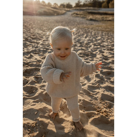 Kleinkind in cremefarbenem Strickoutfit steht am Sandstrand