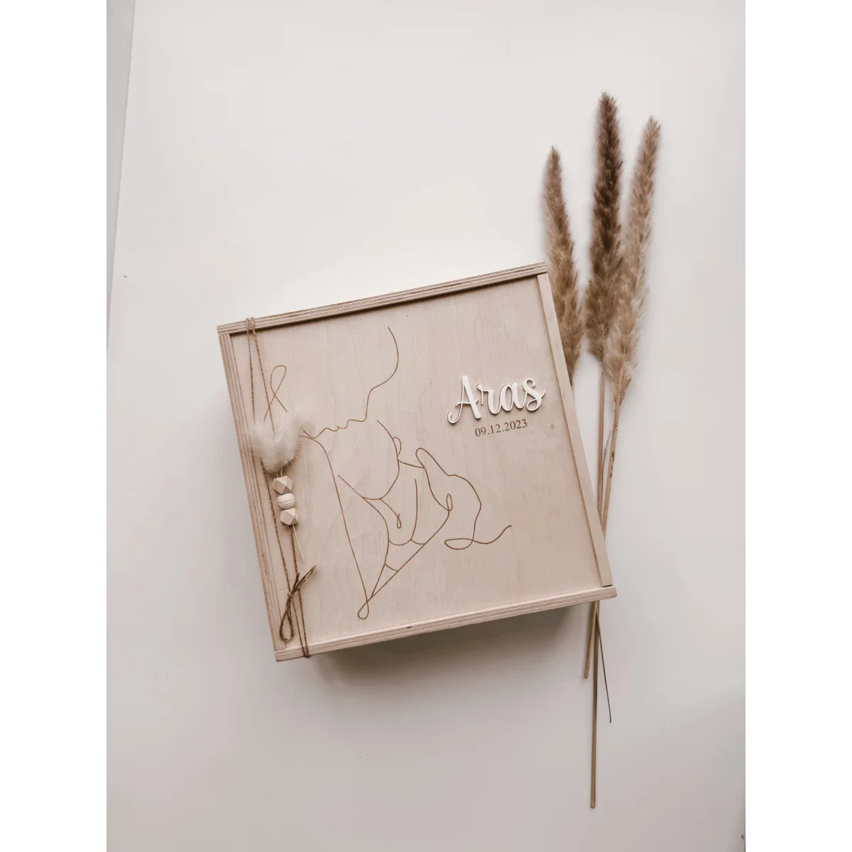 Holzkiste mit Linienzeichnung und Text, umgeben von getrocknetem Pampasgras