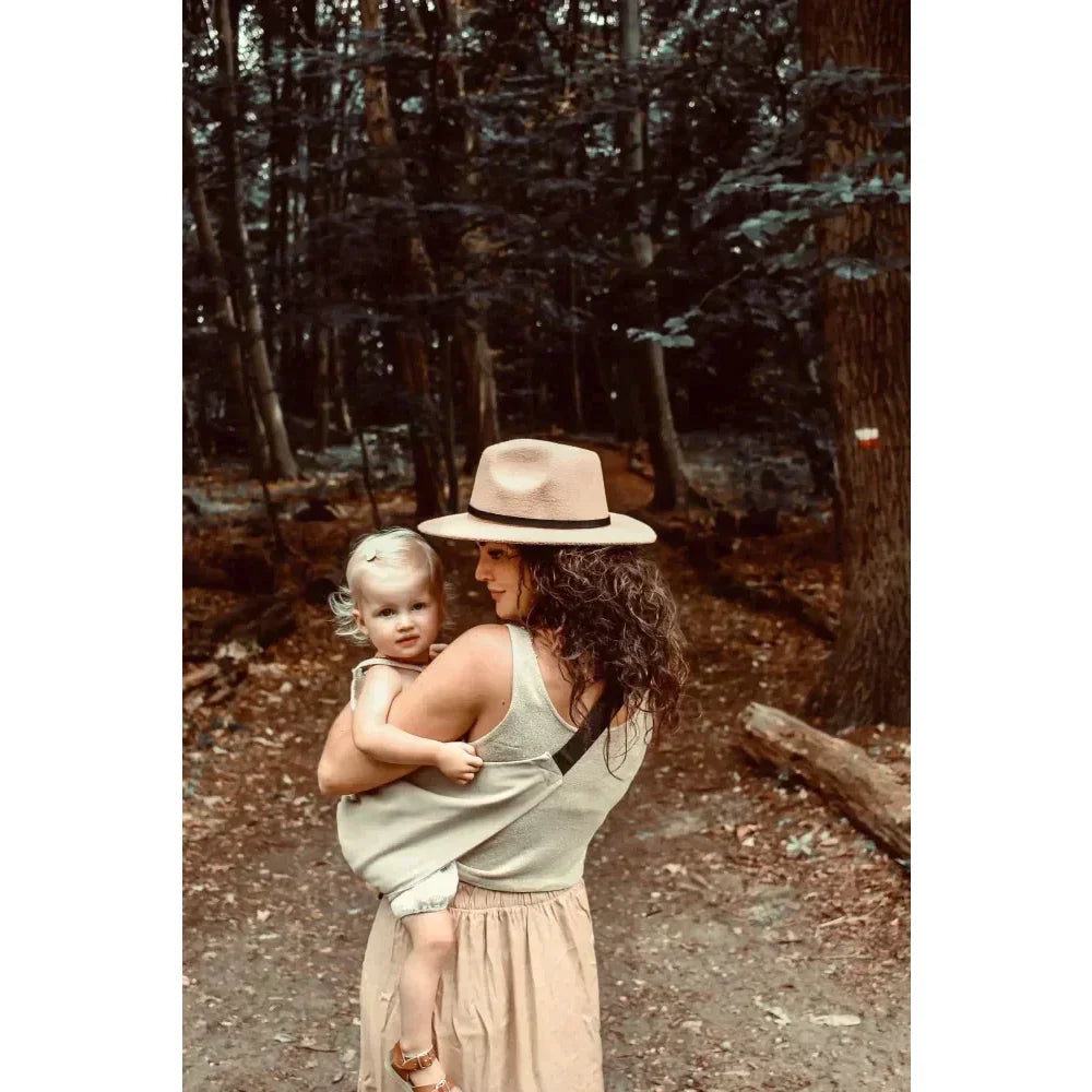 Frau mit Hut und jungem Kind im Wald trägt die Kindertrage in Beige