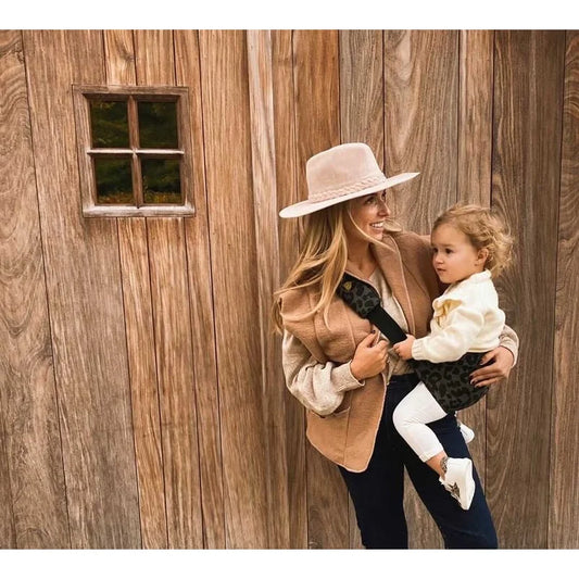 Frau hält Kind und Hund vor einer Holzwand mit Kindertrage Leo grau