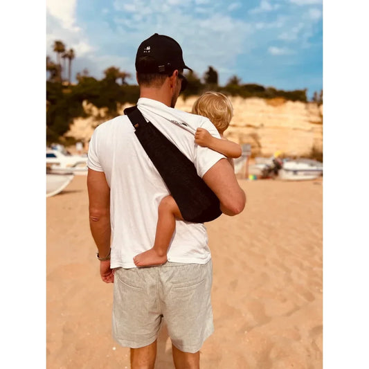 Mann hält ein Kind am Strand mit der Kindertrage in Schwarz