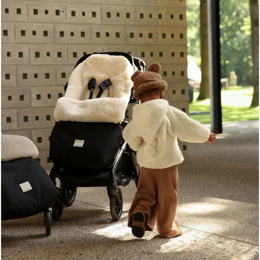 Kind in cremefarbenem Plüschmantel und brauter Mütze schiebt schwarzen Kinderwagen mit weißem Schafsfell-Fußsack Puffmuff Fixed Harness Milk