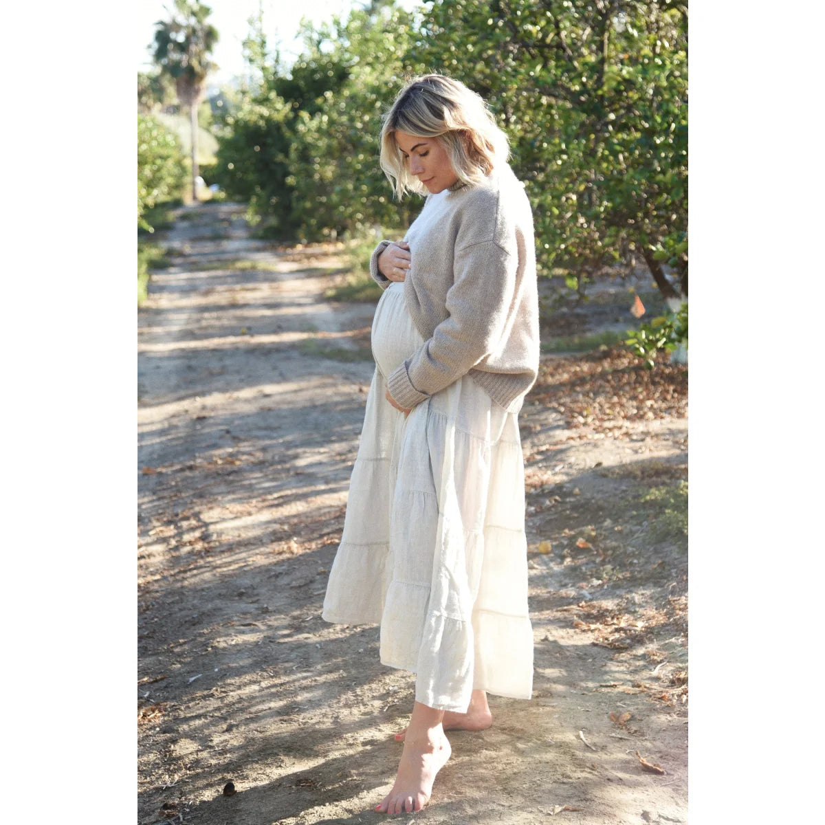 Schwangere Frau in weißem Kleid und beigem Cardigan auf einem Erdweg