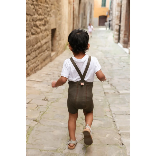 Ein kind trägt eine kurze strumpfhose mit träger in seaweed brown und olivgrüne hosenträger über weißem t-shirt nachhaltige babykleidung schweiz