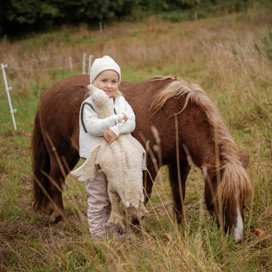 Small child in a white knitted hat beside a brown pony with the Senger Naturwelt Large Cuddly Goose in Linen