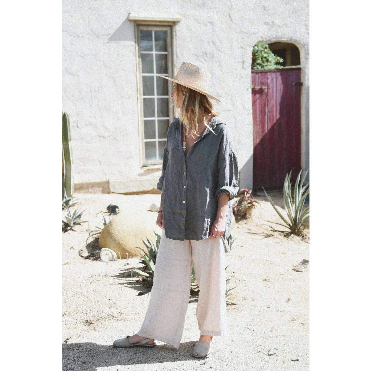 Frau in breitem Hut, dunklem Shirt und hellen weiten Hosen vor weißem Gebäude