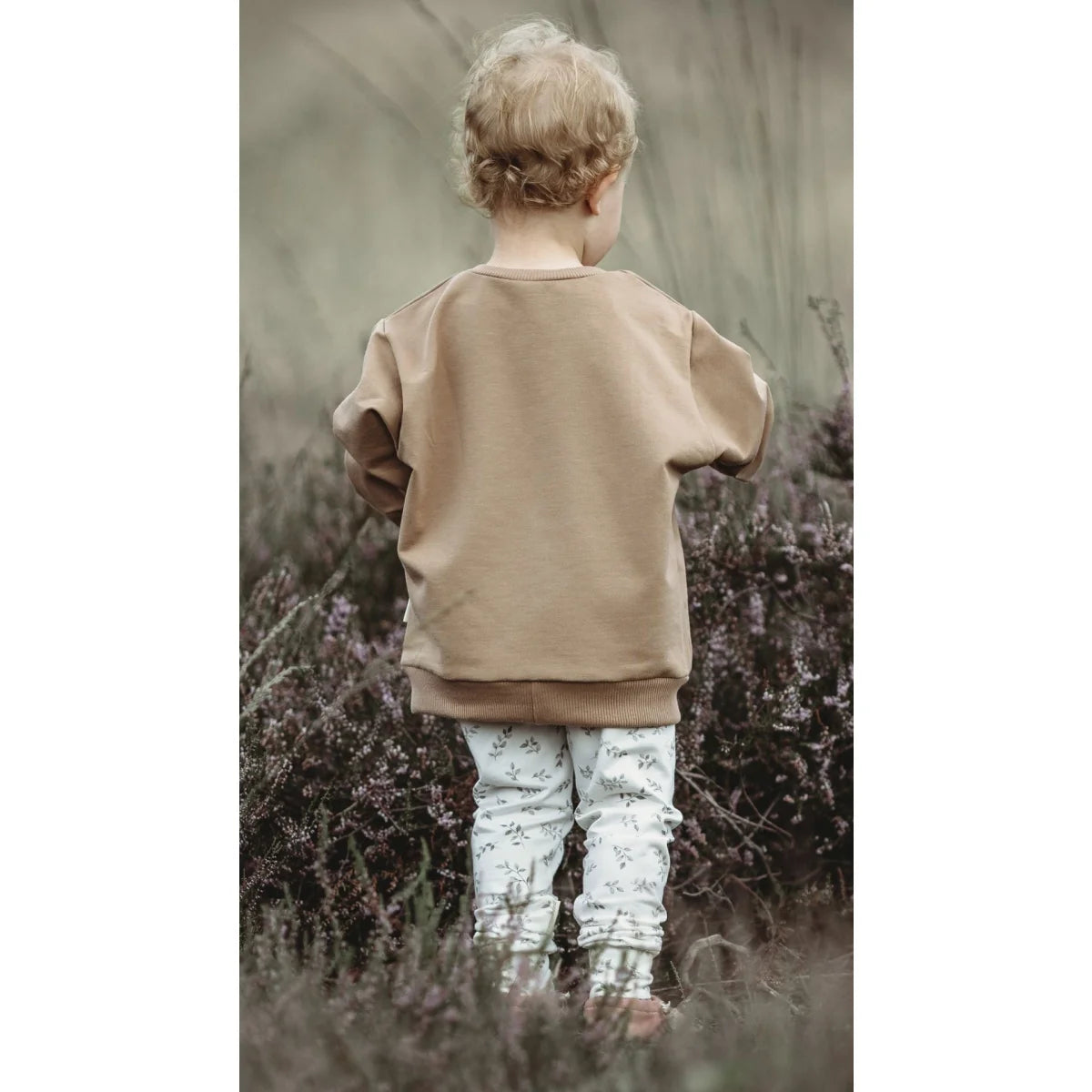Kind mit lockigem blonden Haar in beigem Pullover und gemusterten Hosen, von hinten