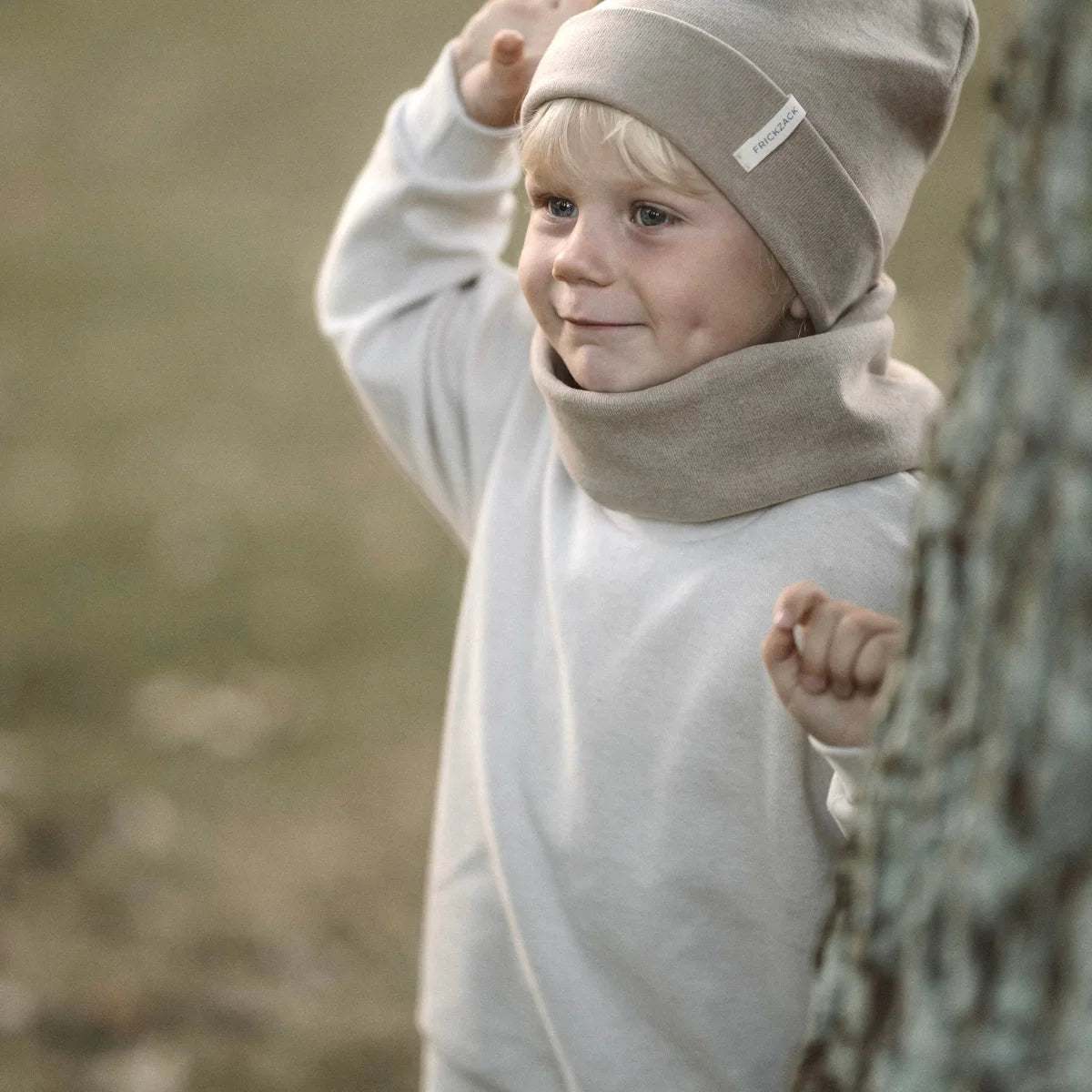 Junger Kind mit beigem Hut und Outfit, schaut hinter Baumstamm hervor