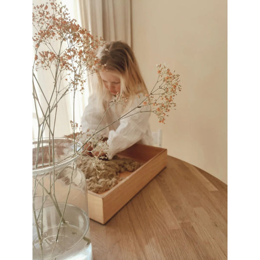 Junges Kind mit langen blonden Haaren im weißen Hemd beim Spielen mit Holztablett voller getrockneter Naturmaterialien