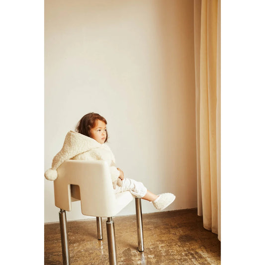 Kind sitzt in einem weißen, skulpturalen Stuhl mit Metallbeinen neben Merino Snugglejacke