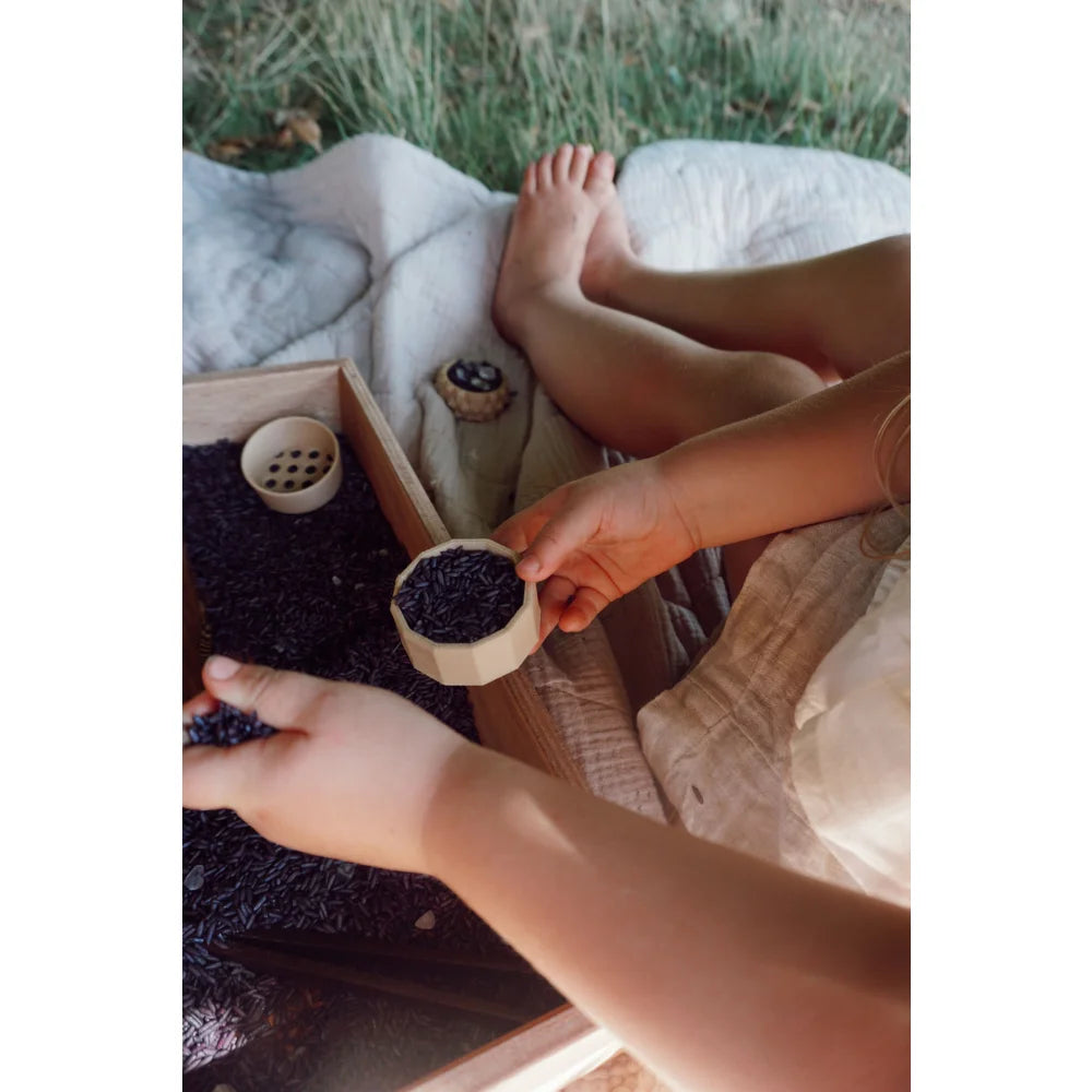 Kinderhände spielen mit dunklen Perlen in einem Holztablett mit kleinen Holzbechern