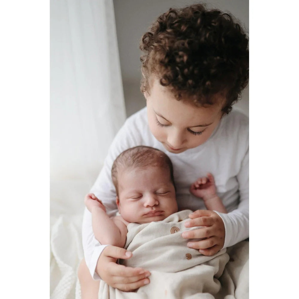 Ein junges Kind hält ein Neugeborenes und schaut es liebevoll an. Mulltuch Rainbow