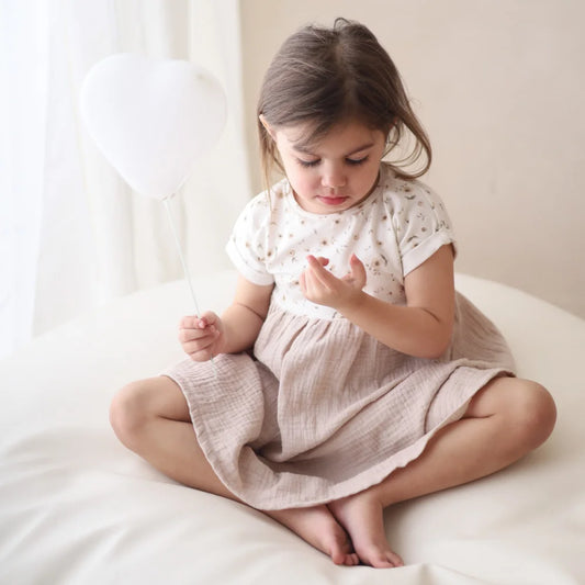 Mädchen im Musselinkleid mit Herzluftballon in blühender Wiese