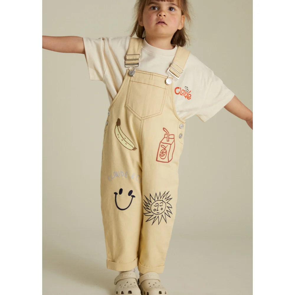 Ein Kind in illustrierten Latzhosen aus dem Piper Breakfast Dungaree Produkt