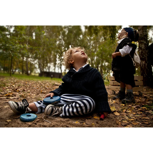 Ein kind mit lockigem blondem haar sitzt im wald auf nachhaltige babykleidung schweiz in schwarz-weiß gestreiften hosen und dunklem strickpullover beim spielen mit dem rainbow round pyramid spielzeug