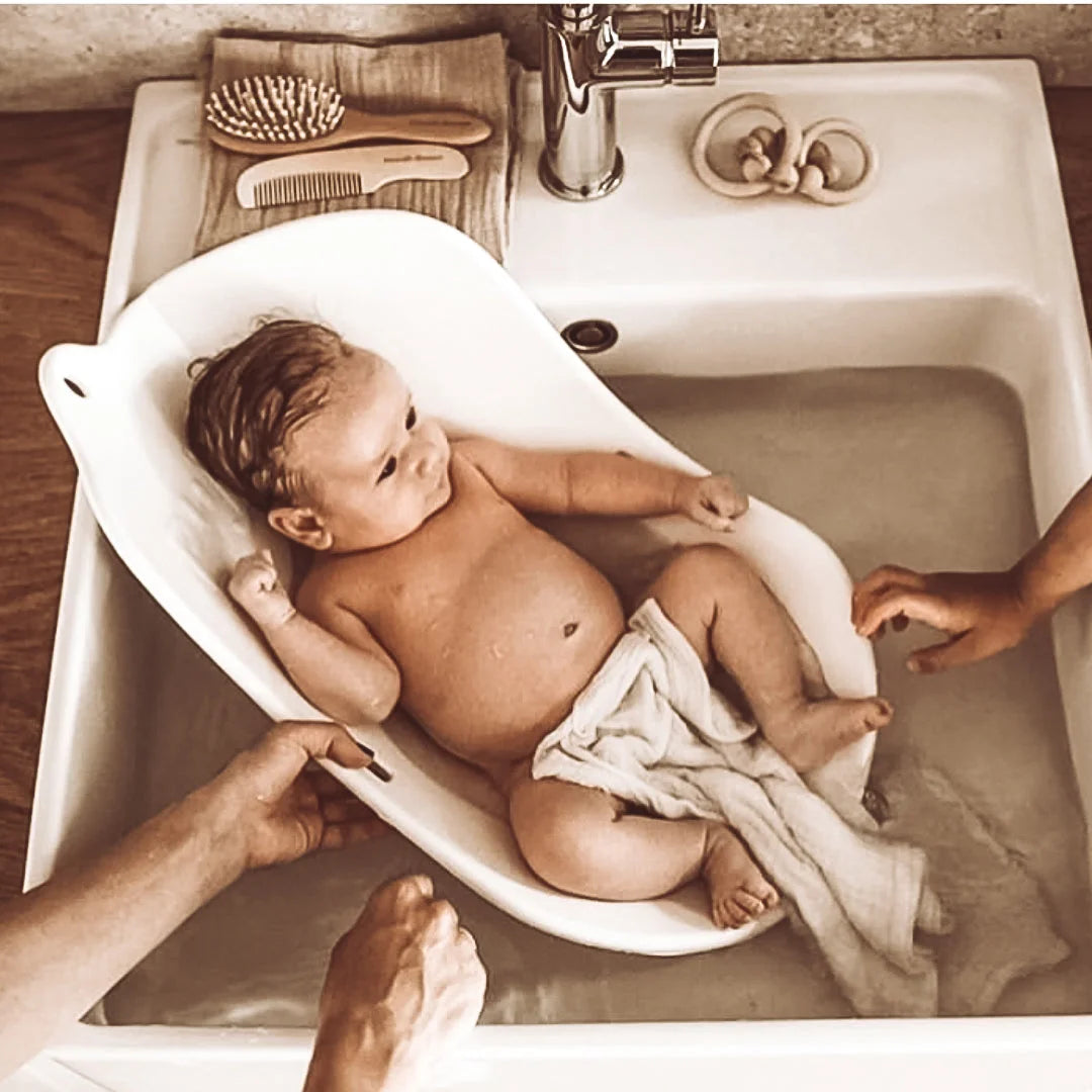 Baby liegt in einem weißen Badeeimer in einer Spüle, Produkt Rassel Wood Naturel