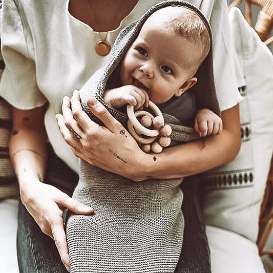 Baby in grauem Strickdecke, von erwachsenen Armen gehalten, Rassel Wood Naturel