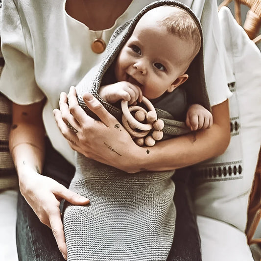 Baby in grauem Strickdecke, von erwachsenen Armen gehalten, Rassel Wood Naturel