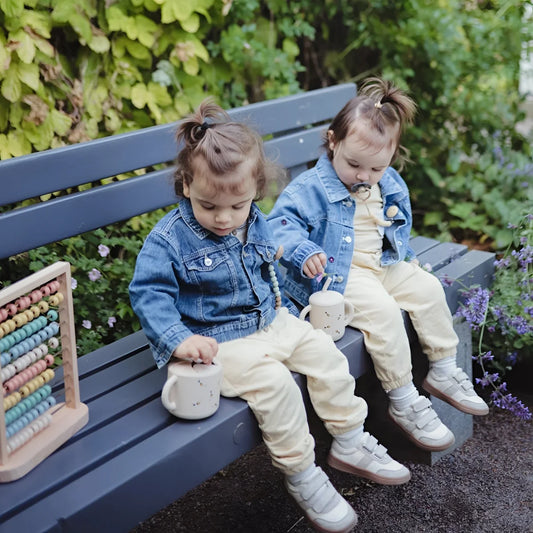 Zwei kleinkinder in blauen jeansjacken und cremefarbenen hosen sitzen mit weißen keramik snack bechern bee auf einer bank