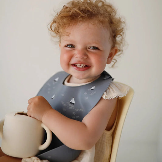 Lächelnder Kleinkind mit lockigem rotem Haar, grauem Lätzchen und Becher