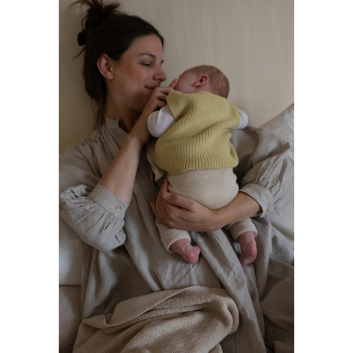 Mutter hält liebevoll ihr Neugeborenes im Strickpullunder Vest Harvey in Hellgelb