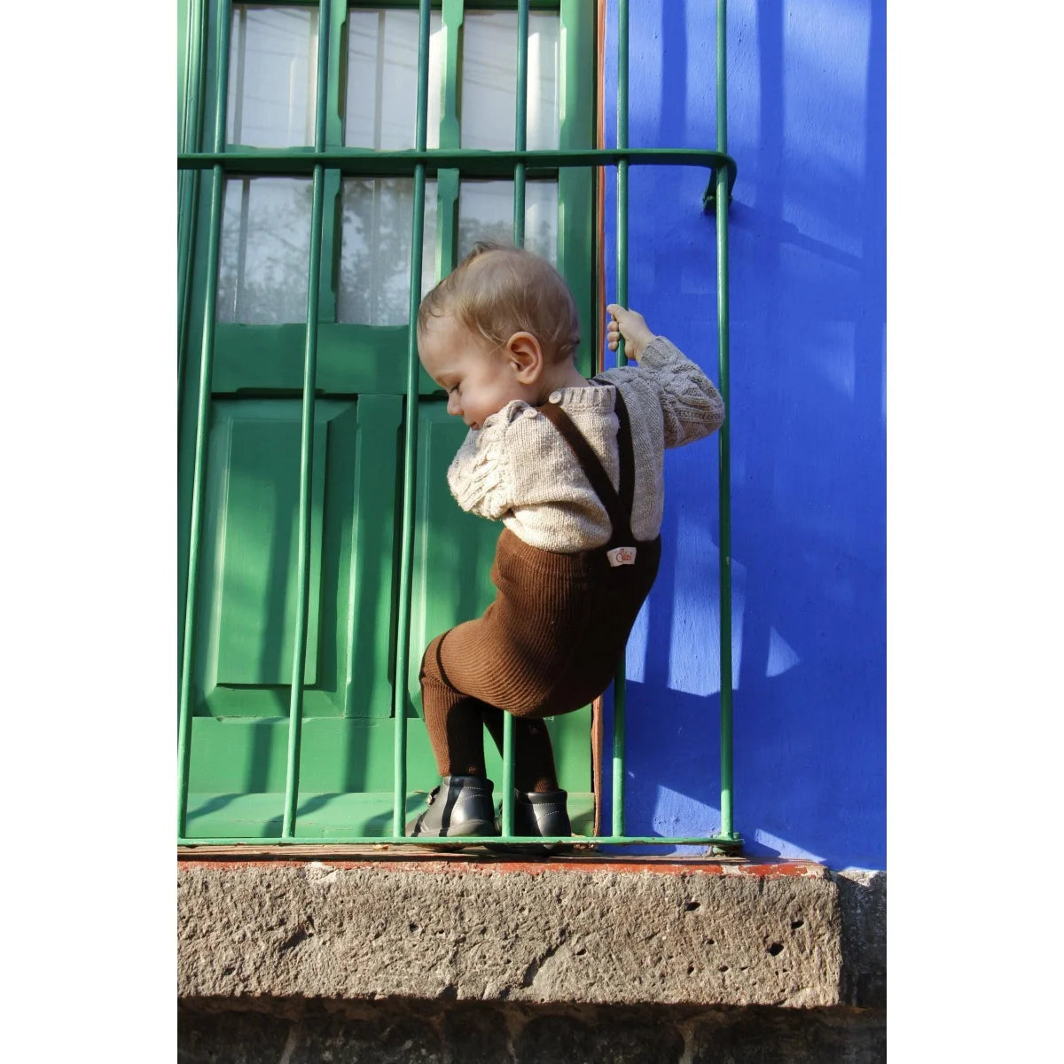 Junges Kind in Hosenträger-Shorts blickt durch grüne Fensterstäbe