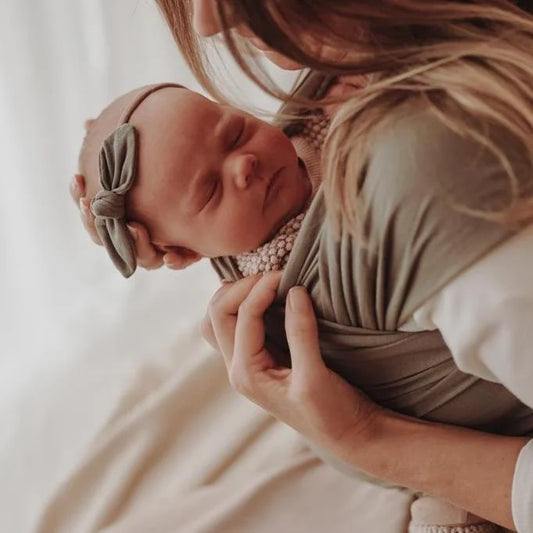 Neugeborenes Baby mit Schleifenhaarband in den Armen einer Person, Tragetuch Dusty Olive