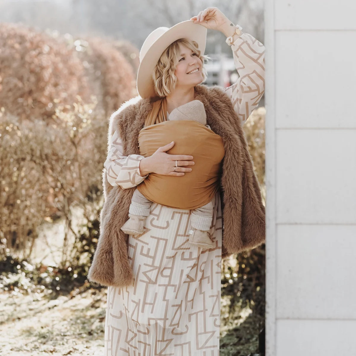 Schwangere Frau in braunem Pelzmantel und gemustertem Boho-Kleid mit Tragetuch Golden Pecan