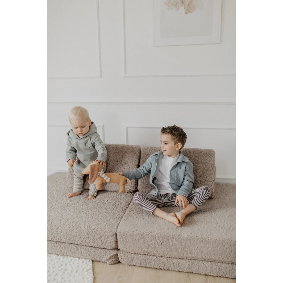 Zwei junge Jungen spielen mit einem Plüschdackel auf einem modularen Sofa in einem grünen Unisex Hoodie