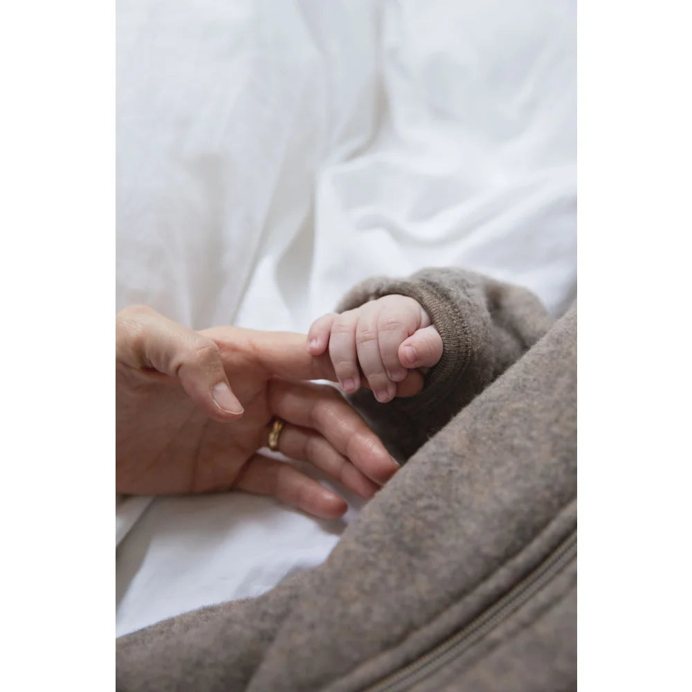 Elternhand mit Goldring hält Babyhand in braunem Wollfleece Zip-Overall mit Kapuze