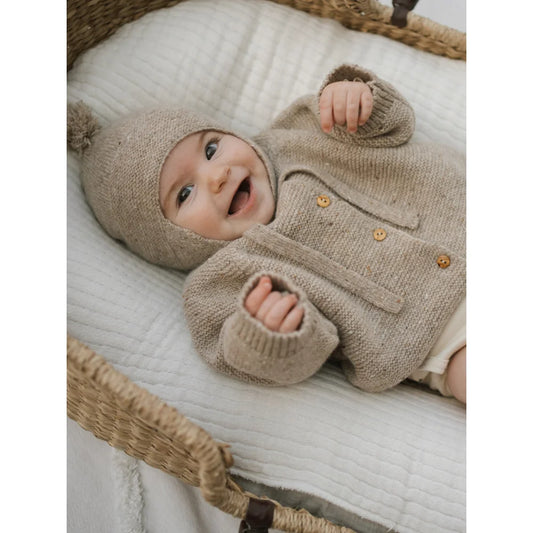 Lächelndes Baby in wolliger Oatmeal Strickjacke mit Pompon und Holzknöpfen