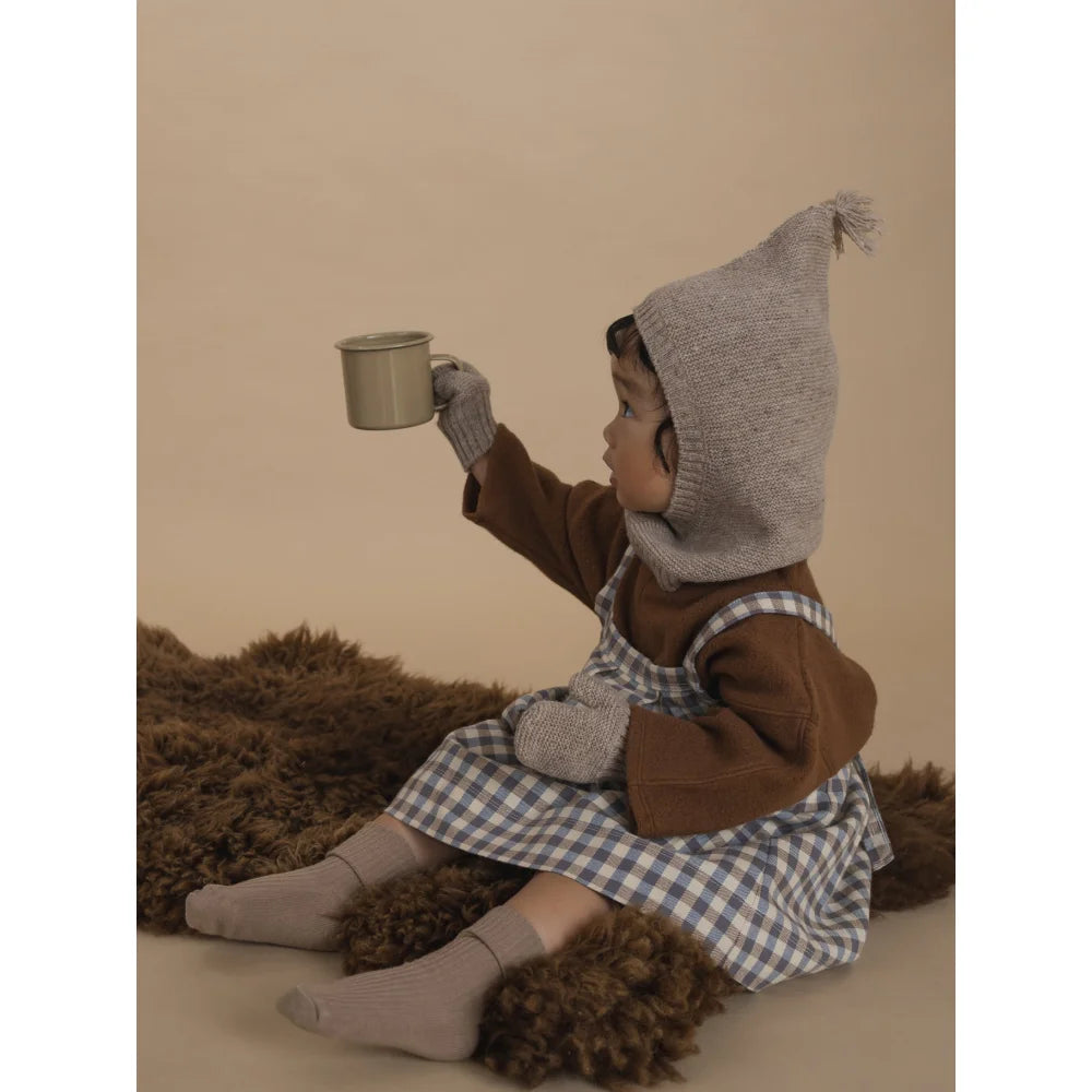 Kleinkind in braunem Hemd, kariertem Kleid, grauem Mützen- und Handschuhset, hält Tasse