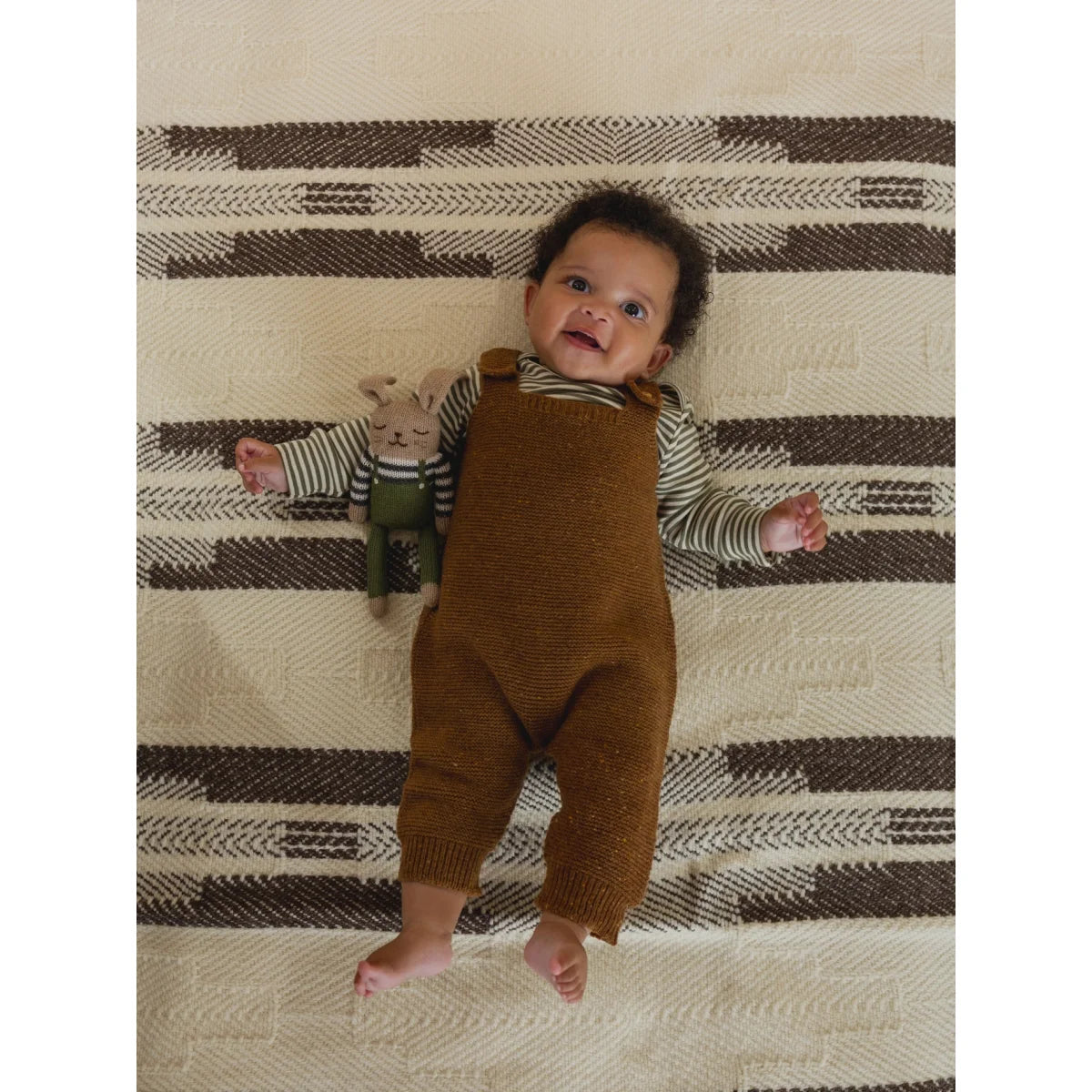 Kleiner Säugling mit dunklen Locken in Wool Salopette Chestnut neben Plüschhasen