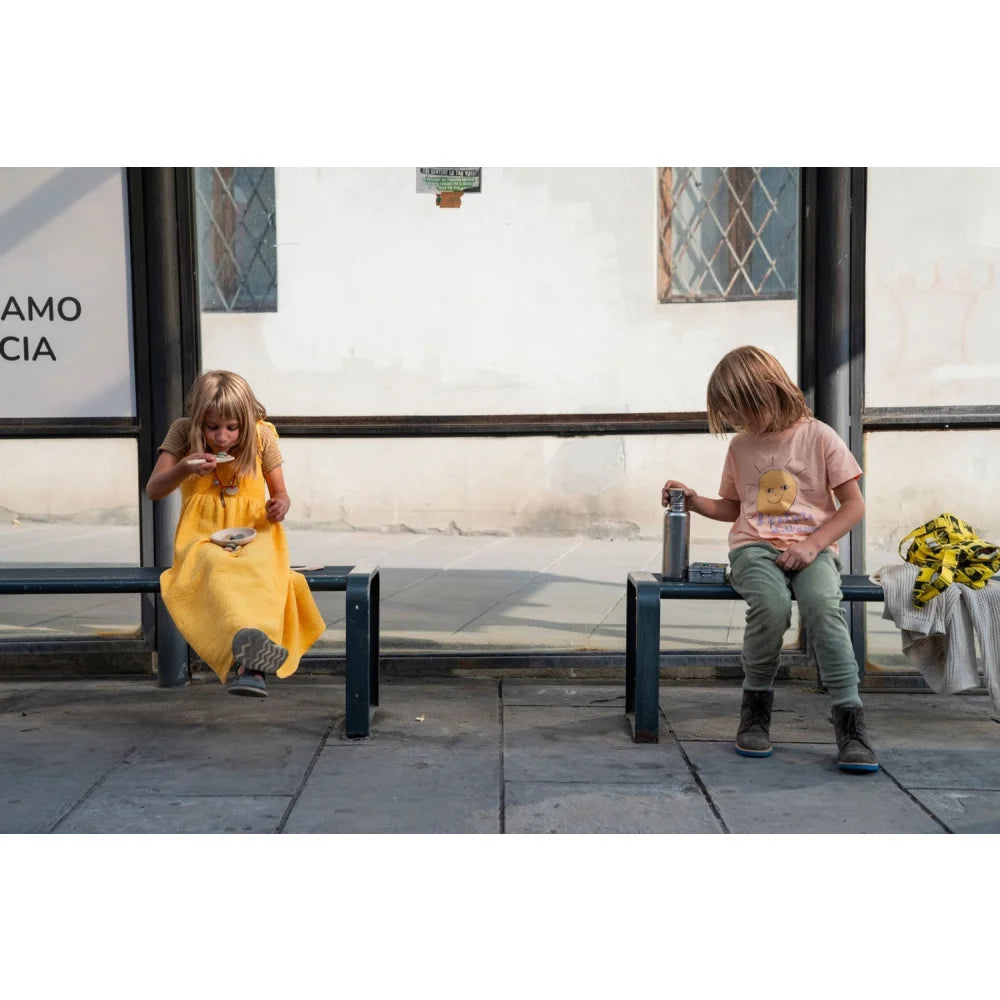 Kind in gelbem Kleid sitzt auf dunkler Bank an Bushaltestelle Yummy