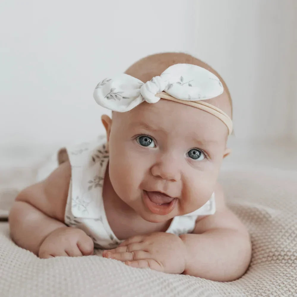Ein glückliches Baby in einem weißen Haarband und floralem Outfit aus der NEWBORN-Kollektion.