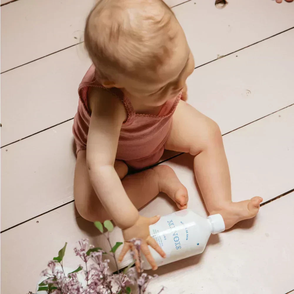 Baby in rosa Strampler spielt mit weißer Flasche und lila Blumen bei Natürliche Babypflege